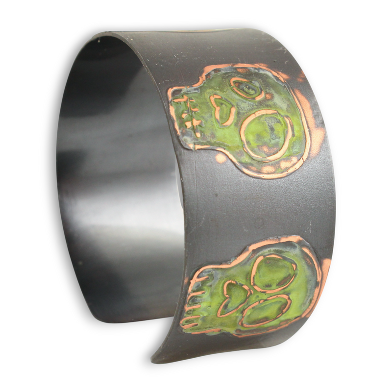 Oxidized metal cuff bracelet with green coloring over skull designs on a white background.