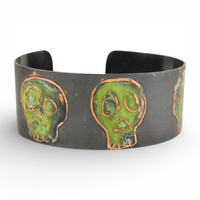 Oxidized metal cuff bracelet with green coloring over skull designs on a white background.