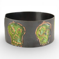 Oxidized metal cuff bracelet with green coloring over skull designs on a white background.