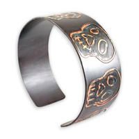 Metallic cuff bracelet with intricate blue copper skull designs on a white background