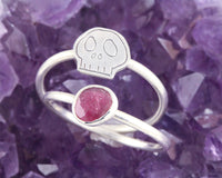 Two silvers ring together, featuring one with a pink gemstone and the other with an engraved skull motif, displayed on a bed of purple amethyst crystals.