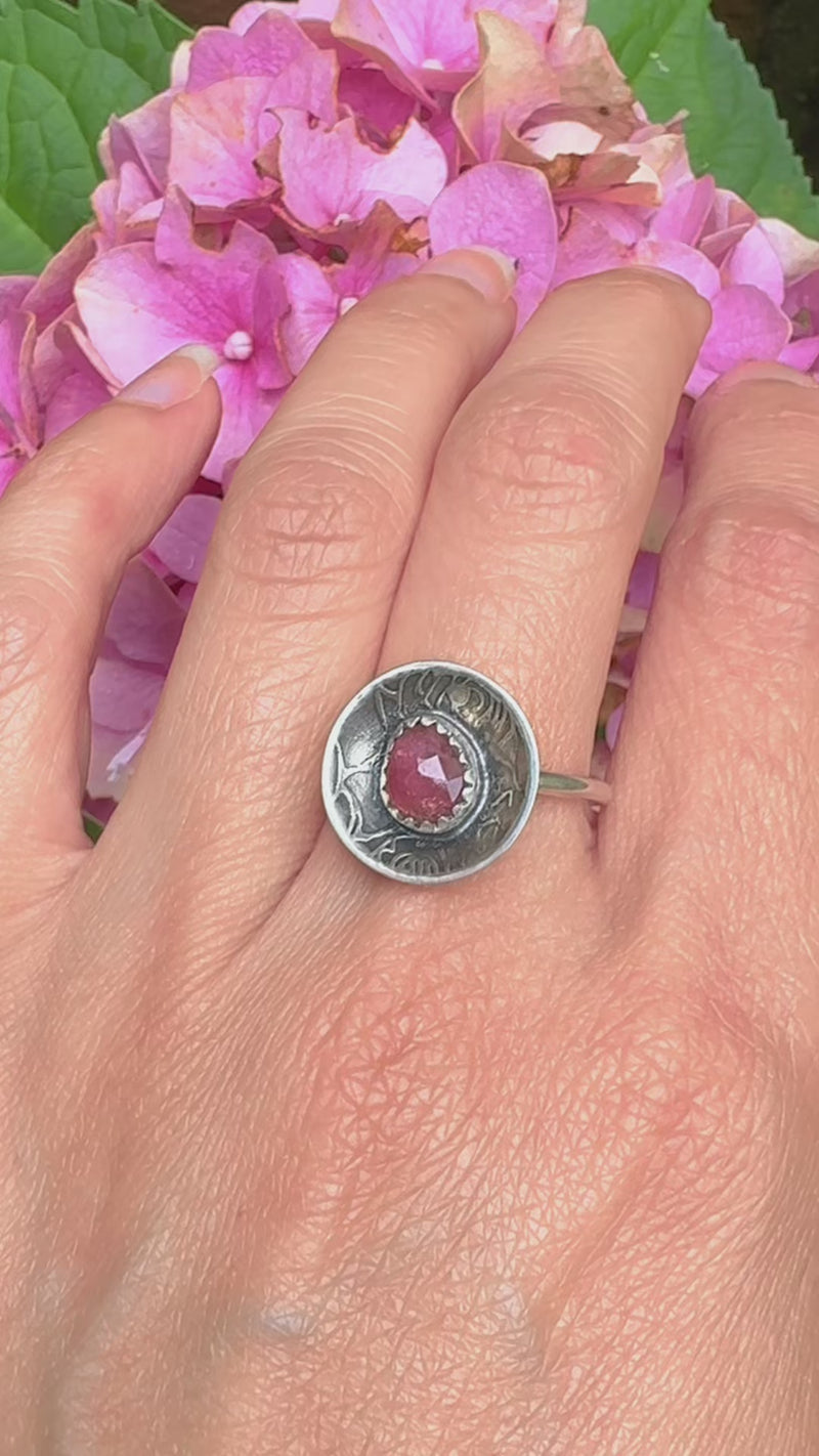 A sterling silver ring featuring a round, rose pattern textured dome set with a faceted pinkish-red gemstone, modeled on the maker that made it.