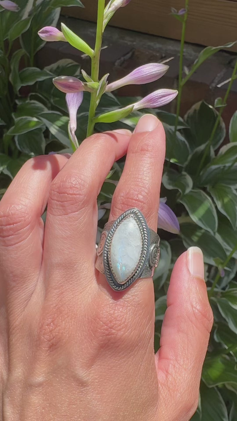 Video of a hand modeling - A wide silver ring featuring a large oval diamond cabochon moonstone with a soft gray-blue iridescent sheen, set in a textured band with a decorative skull motif.
