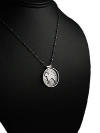 A round silver-toned metal pendant featuring a raised dog silhouette over an embossed pattern of bones, shown on a black display bust.