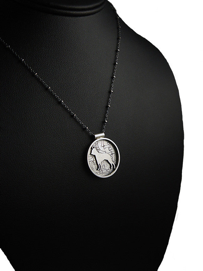 A round silver-toned metal pendant featuring a raised dog silhouette over an embossed pattern of bones, shown on a black display bust.